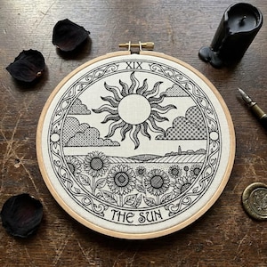 May include: A circular embroidery hoop containing a white fabric panel with a black line art illustration of a tarot card. The design features a large sun with wavy rays, clouds, a field of sunflowers, and the text XIX and THE SUN.