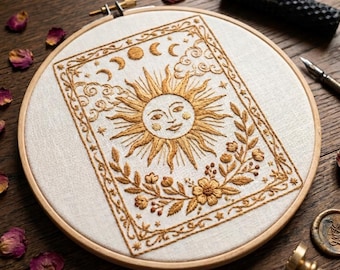May include: A circular wooden embroidery hoop containing a white fabric panel with gold thread embroidery. The design features a central sun face with rays, surrounded by moon phases, clouds, stars, and floral vines within a rectangular border.