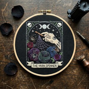 Könnte beinhalten: Ein runder hölzerner Stickrahmen mit schwarzem Stoff und einem handgestickten Motiv. Die Stickerei zeigt eine Hand, die eine Häkelnadel hält, ein Spinnennetz, drei Wollknäuel, zwei Rosen, zwei Totenköpfe und den Text THE YARN SPINNER.