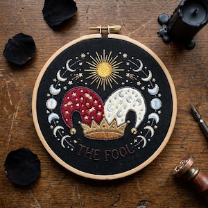 May include: A circular embroidery hoop containing black fabric with a hand-stitched design of a jester hat in red and white, a gold crown, a yellow sun, and a lunar cycle. The text THE FOOL is stitched in brown thread at the bottom centre.