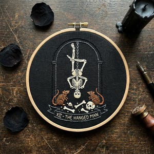 Op de afbeelding: Een ronde houten borduurring met zwarte stof en wit borduurwerk van een skelet dat ondersteboven aan een ketting hangt. Twee bruine muizen zitten onderaan bij verspreide botten en een schedel. De tekst XII - THE HANGED MAN is eronder geborduurd.