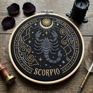 Könnte beinhalten: Ein runder Stickrahmen mit schwarzem Stoff, der in der Mitte einen grauen Skorpion zeigt, umgeben von goldenen und silbernen Himmelsmustern, Sternen und Sternbildern. Das Wort SCORPIO ist unten im Design in Gold gestickt.