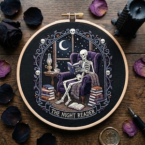 May include: An embroidery hoop containing a black fabric panel with a detailed stitched scene of a skeleton sitting in a purple armchair reading a book titled THE NIGHT READER. The design includes stacks of books, a lamp, and a crescent moon.
