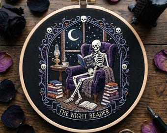 May include: A circular embroidery hoop containing a black fabric panel with a detailed thread-stitched scene of a skeleton sitting in a purple armchair reading a book titled The Night Reader, surrounded by stacks of books and skulls.