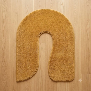 May include: A mustard yellow, U-shaped bath mat with a textured, looped pile surface. The mat features a rounded top edge and a central cutout, creating a distinct arch shape with finished, matching fabric edges along the entire perimeter.