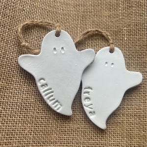 May include: Two white ghost-shaped ornaments made of a smooth material, each featuring two small indented eyes and a name stamped into the surface. One ghost is stamped with the name callum and the other with freya, both hanging from twine loops.