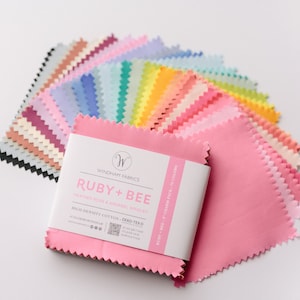 May include: A stack of colorful fabric swatches in various shades of pink, blue, green, yellow, and orange. The swatches are arranged in a rainbow pattern. The fabric is labeled "Ruby + Bee" by Windham Fabrics. High density cotton, Oeko-Tex certified.