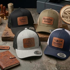 May include: Four baseball caps with leather patches featuring the text Aufbein Truces, Northline Atelier, Adventure Co., and Saguenay Brass Byd. The caps are black, olive green, heather grey, and navy blue with mesh backs and curved brims.