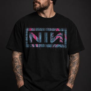Op de afbeelding: Een zwart T-shirt met korte mouwen en een rechthoekige afbeelding op de borst. De afbeelding bevat de letters NIИ in een vet, gestileerd lettertype, geplaatst tegen een achtergrond van wervelende blauwe en roze marmerpatronen met een textuurrand.