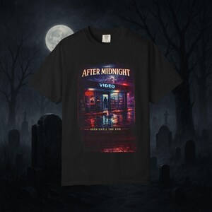 以下が含まれることがあります： 夜のビデオ店を描いたグラフィックプリント付きの黒い半袖Tシャツ。テキストには「AFTER MIDNIGHT」および「OPEN UNTIL THE END」と書かれています。画像には、ネオンサイン、濡れた舗装、暗く嵐のような空を背景にした店舗の正面が描かれています。