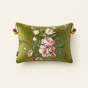 May include: A rectangular olive green velvet cushion featuring an embroidered floral branch design with pink and white blossoms. The cushion is accented with small, round, multi-coloured fabric tassels attached to each of the two side corners.