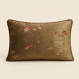 May include: A rectangular throw pillow in a muted olive green shade featuring delicate embroidered floral vines with pink and red rose blossoms. The pillow is finished with a thin, dark brown piped edge that frames the entire rectangular shape.