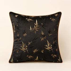 Peut inclure: Un coussin carré doté d'une base en tissu noir avec un motif brodé de feuilles de bambou en fil doré métallique et foncé. Le coussin est fini avec un passepoil fin et uni de couleur marron le long des quatre bords, créant une bordure définie.