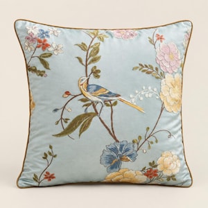Peut inclure: Un coussin carré à fond bleu clair orné d'une broderie complexe représentant un oiseau perché sur une branche, entouré de fleurs variées dans des tons de rose, jaune, bleu et blanc. Le coussin est fini avec un passepoil marron.