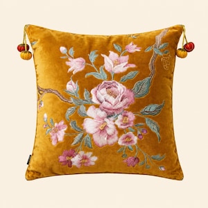 Peut inclure: Un coussin carré en velours jaune moutarde orné d'un motif floral brodé complexe avec des fleurs roses et blanches, des feuilles vertes et des branches brunes. Le coussin est agrémenté de petits pompons rouges et jaunes aux coins supérieurs.
