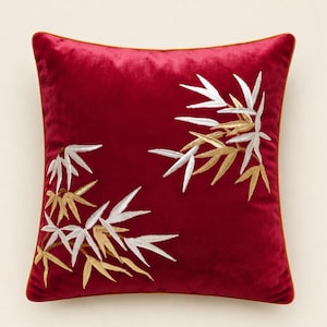 May include: A square velvet throw pillow in a deep red color featuring embroidered bamboo leaf patterns in metallic gold and silver thread. The pillow is finished with a thin orange piping along the edges, creating a distinct border for the design.