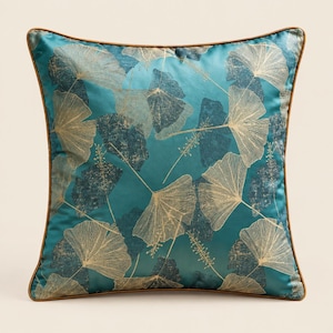 Peut inclure: Un coussin carré en tissu satiné bleu sarcelle orné d'un motif répétitif de feuilles de ginkgo dorées et de formes de feuilles texturées bleu sarcelle foncé. Le coussin est bordé d'un fin passepoil marron uni sur les quatre côtés.