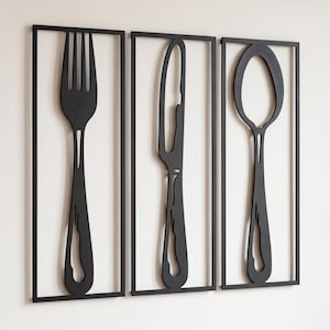 May include: A set of three rectangular black metal wall art pieces, each featuring a cutout silhouette of a single piece of cutlery: a fork, a knife, and a spoon. The frames are thin and rectangular, enclosing the black utensil shapes.