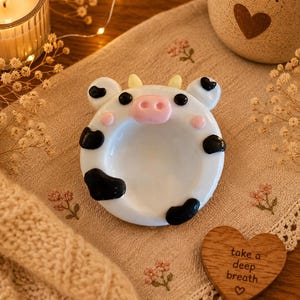 May include: A round white ceramic trinket dish shaped like a cow face with black spots, small yellow horns, and a pink snout. The dish features a smooth, glossy finish and a concave center, designed with a whimsical animal-inspired aesthetic.