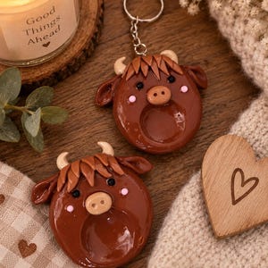 May include: Two brown Highland cow-shaped ornaments with glossy finishes, featuring cream-colored horns, dark eyes, and pink cheeks. One ornament is attached to a silver metal keychain ring with a chain, while the other is a standalone circular piece.