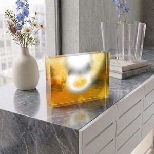 Puede incluir: Un bloque rectangular de acrílico con un diseño abstracto en tonos vibrantes de amarillo, naranja y dorado. La pieza tiene una superficie lisa y pulida que crea un efecto brillante con un reflejo de luz circular blanco en el centro.
