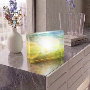 May include: A rectangular acrylic block featuring a printed landscape scene of a bright sun shining over a green field. The clear block displays a vibrant yellow and green colour palette with a central white light effect, creating a luminous appearance.