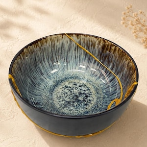 May include: A round ceramic bowl featuring a reactive glaze with a deep blue and white speckled interior that transitions to a dark brown rim. A distinct, thin, golden-yellow line runs diagonally across the interior surface of the bowl.