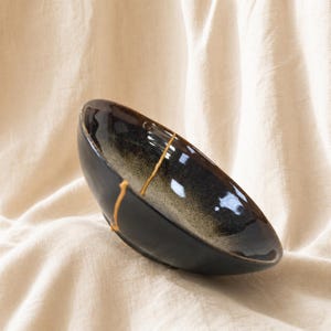 May include: A ceramic bowl with a dark, glossy finish that transitions into a lighter, speckled grey centre. The bowl features a prominent, jagged crack running across its surface, which is filled with a bright, metallic gold-coloured material.