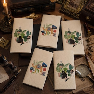 Puede incluir: Cinco cuadernos rectangulares de papel con una cubierta texturizada de color blanco roto. Tres cuadernos presentan un gato negro sentado entre plantas verdes en macetas y cactus. Dos cuadernos muestran un camello beige rodeado de flores rojas y azules.