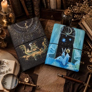Puede incluir: Dos diarios rectangulares con bandas elásticas. El diario de la izquierda es negro con patrones celestiales dorados y una ilustración de un barco. El de la derecha es azul con una escena de ventana con un conejo y una figura.