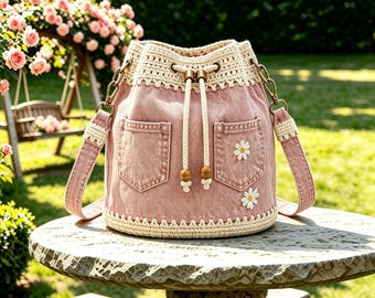 May include: A dusty pink denim bucket bag featuring a cream-colored crocheted top border and base. The bag includes two front pockets, a drawstring closure with wooden beads, and a matching pink denim shoulder strap with cream crochet accents.