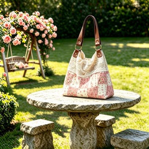 Op de afbeelding: Een hobo-handtas met een patchworkontwerp van roze en witte bloemrijke stoffen vierkanten. Het bovenste gedeelte is versierd met crèmekleurige kanten randen en de tas heeft twee bruine lederen schouderbanden bevestigd met goudkleurige ringen.
