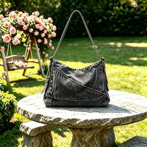 May include: A dark gray denim shoulder bag featuring a slouchy silhouette, a single top handle, and a diagonal front flap secured with metallic buttons. The bag is constructed from washed fabric with visible seams and a structured, curved base design.
