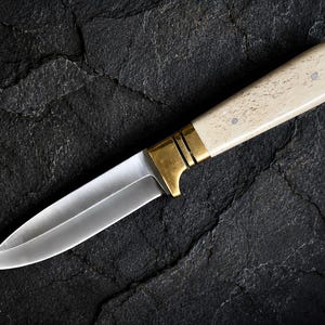 May include: A fixed-blade knife featuring a polished silver-toned steel blade, a brass bolster with two horizontal grooves, and a smooth, cream-colored handle with a speckled texture secured by two small metal pins visible on the side.