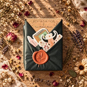 May include: A rectangular package wrapped in black paper, sealed with a terracotta-colored wax seal featuring a leaf design. The package contains a card with the text Your Ritual Awaits and two stickers, one of a book and one of a bull skull.
