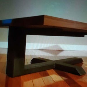 Könnte beinhalten: Ein Couchtisch mit einer rechteckigen Holzplatte in glatter, hellbrauner Ausführung. Der Tisch wird von einem schwarzen Metallgestell getragen, das aus einem einzelnen vertikalen Pfosten besteht, der mit einem breiten, kreuzförmigen Fuß verbunden ist.
