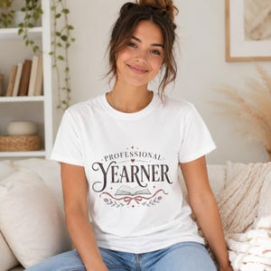 Può includere: Una t-shirt bianca a maniche corte con il testo Professional Yearner in un carattere serif scuro. Il design include un piccolo cuore, un grafico di un libro aperto ed elementi floreali decorativi con un nastro in inchiostro rosso e nero.