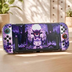 Peut inclure: Une console de jeu portable dotée d'un habillage violet et noir avec un motif représentant le personnage Gengar. Le design illustre une scène de cimetière effrayante avec une pleine lune, des grilles en fer et des yeux rouges brillants.