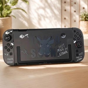 May include: A black handheld gaming console featuring a central graphic of a gray Pikachu character. The device includes white text that reads TOKYO SOON! and HOT MADE IN CHINA, along with small icons of lightning bolts and Poké Balls on the controllers.