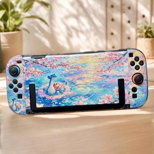 Könnte beinhalten: Eine tragbare Spielkonsole mit einer dekorativen Folie, die ein impressionistisches Gemälde einer blauen Kreatur im Wasser zeigt, umgeben von rosa Kirschblütenzweigen. Das Gerät verfügt über schwarze Tasten, Joysticks und einen Ständer.