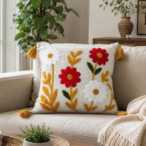 Op de afbeelding: Een vierkant sierkussen met een lichtbeige stoffen basis voorzien van een getuft bloemenpatroon. Het ontwerp bevat rode en witte bloemen met gele en groene stengels en bladeren. Aan de hoeken van het kussen zijn vier gele kwastjes bevestigd.