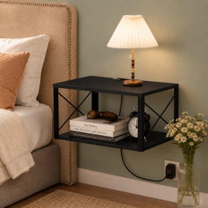 May include: A black floating wall-mounted nightstand with an X-shaped side frame. The top surface holds a pleated white lamp with a gold base. The lower shelf contains two stacked books titled Jean Matisse and Picasso the Creations, and a black alarm clock.