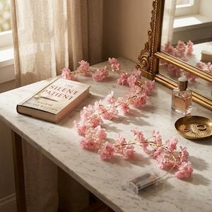 May include: A string of artificial pink cherry blossom flowers with integrated LED lights, a clear plastic battery pack, a hardback book titled The Silent Patient, a glass perfume bottle, and a small gold-toned metal dish on a marble surface.