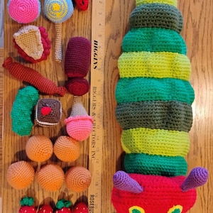 May include: A large crocheted caterpillar toy with a red head, yellow eyes, and purple antennae, accompanied by various crocheted food items including fruit, cheese, and sweets, arranged next to a wooden ruler measuring up to 26 inches.