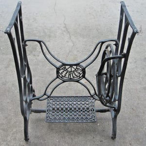 May include: A black cast iron sewing machine base featuring an ornate, curved frame with four legs and a central oval medallion embossed with the word PEERLESS. The structure includes a rectangular foot treadle with a repeating circular pattern.