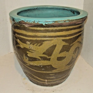May include: A large, round ceramic planter featuring a dark brown body with a prominent, lighter-colored dragon motif wrapped around the exterior. The wide, circular rim is finished in a contrasting bright turquoise glaze with visible wear.