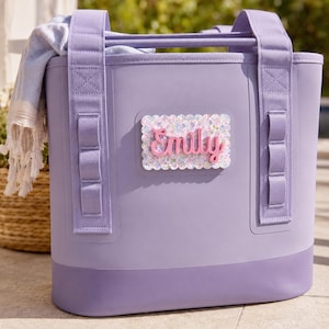 May include: A lavender tote bag with two sturdy handles and vertical webbing straps. A rectangular patch with a scalloped edge and a floral pattern is centered on the front, featuring the name Emily in raised, pink, cursive script lettering.