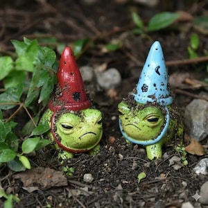 May include: Two small ceramic frog figurines partially covered in dirt. One frog wears a pointed red hat with black spots, and the other wears a light blue pointed hat featuring a small black crescent moon and flower design on the surface.