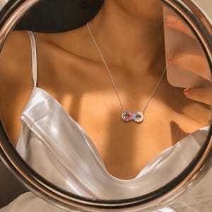 Könnte beinhalten: Eine silberfarbene Halskette mit einem Unendlichkeitssymbol-Anhänger, der mit kleinen, bunten Steinen in Blau-, Rosa- und Weißtönen besetzt ist. Der Anhänger hängt an einer zarten silberfarbenen Kette und liegt auf einem weißen Satin-Kleidungsstück.