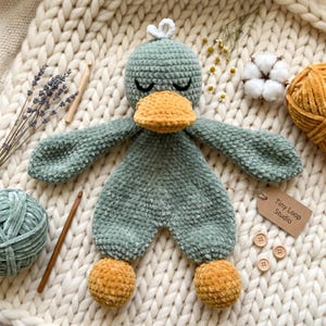 Puede incluir: Un juguete de peluche tejido a ganchillo en color verde salvia con forma de pato, con pico y pies de color amarillo mostaza. El juguete tiene los ojos cerrados, un pequeño lazo blanco en la parte superior y una etiqueta Tiny Loop Studio.